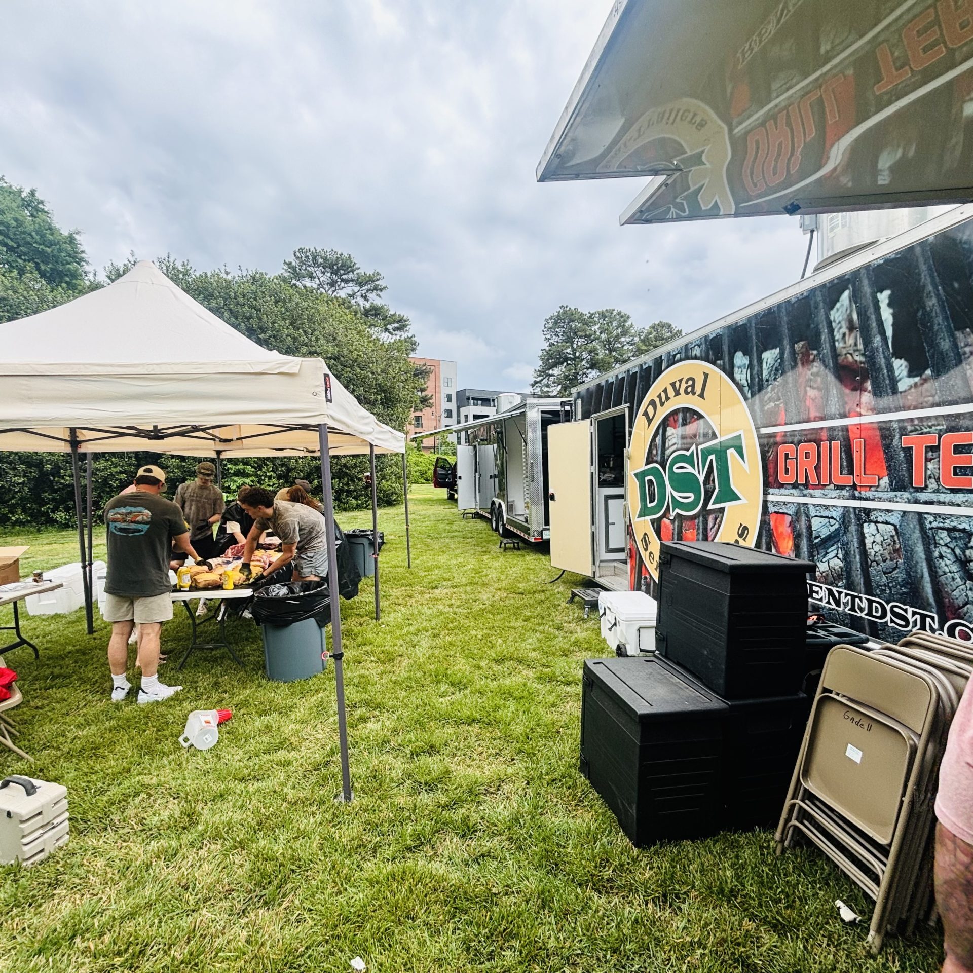 DST employees prepare food and supplies under a canopy tent beside a mobile grilling trailer during a community outreach event.